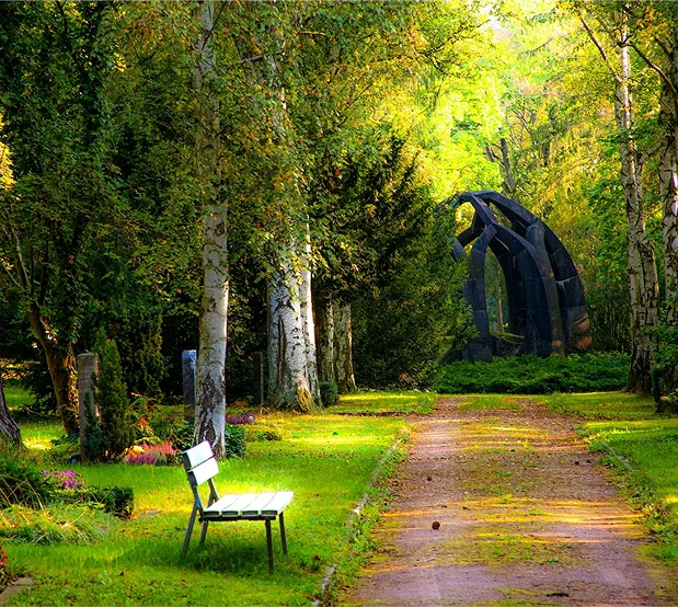 truma-therapy8 A serene garden path with a white bench, leading towards a large, dark abstract sculpture amidst trees.